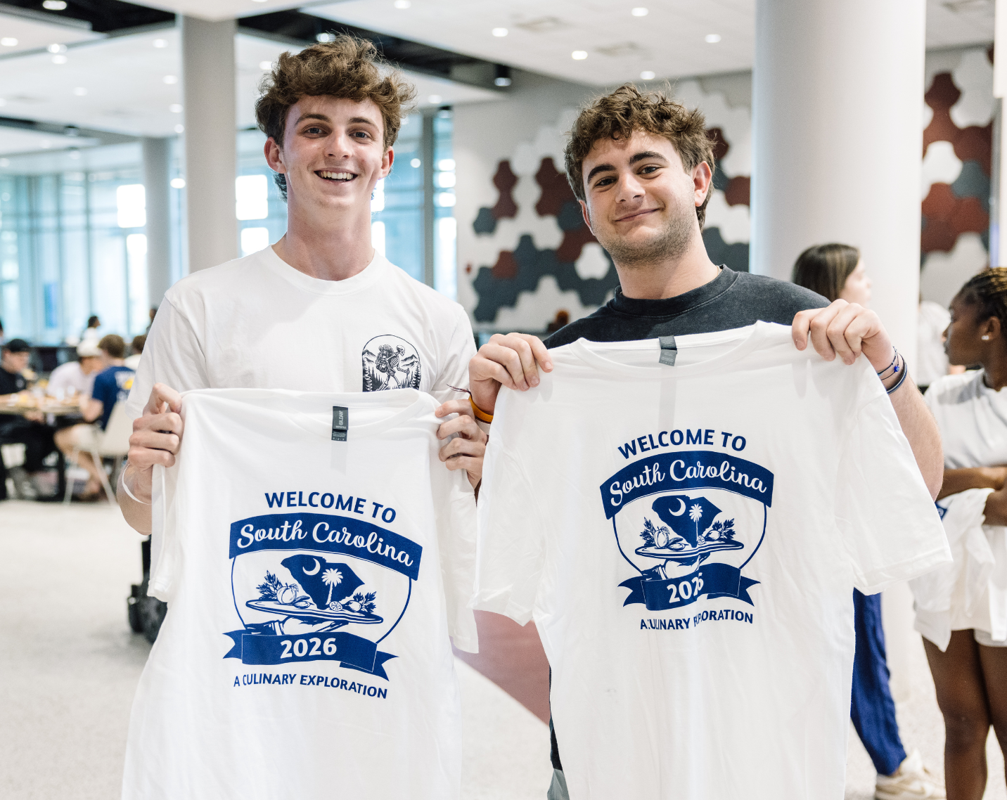 Two students holding up two "Welcome to South Carolina" event t-shirts.